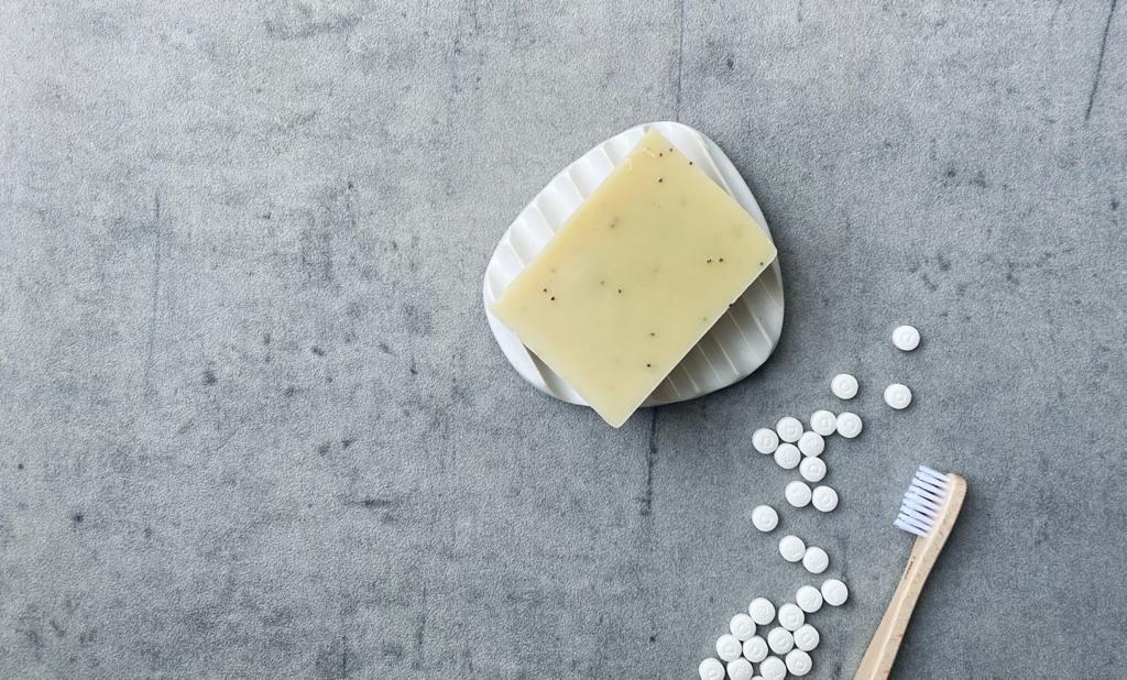 kaikua Mohnsamen und Litsea Cubeba Seife auf weißer Seifenschale. Bambuszahnbürste und Zahnputztabletten unten rechts im Bild